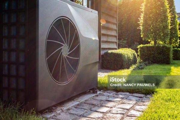 Close-up of black full inverter heat pump outside in the garden, near wooden pool house on a sunny day. Lens flare on the image.